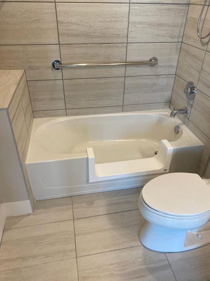 Modern bathroom with white soaking tub, chrome grab bar, beige tile walls, and white toilet